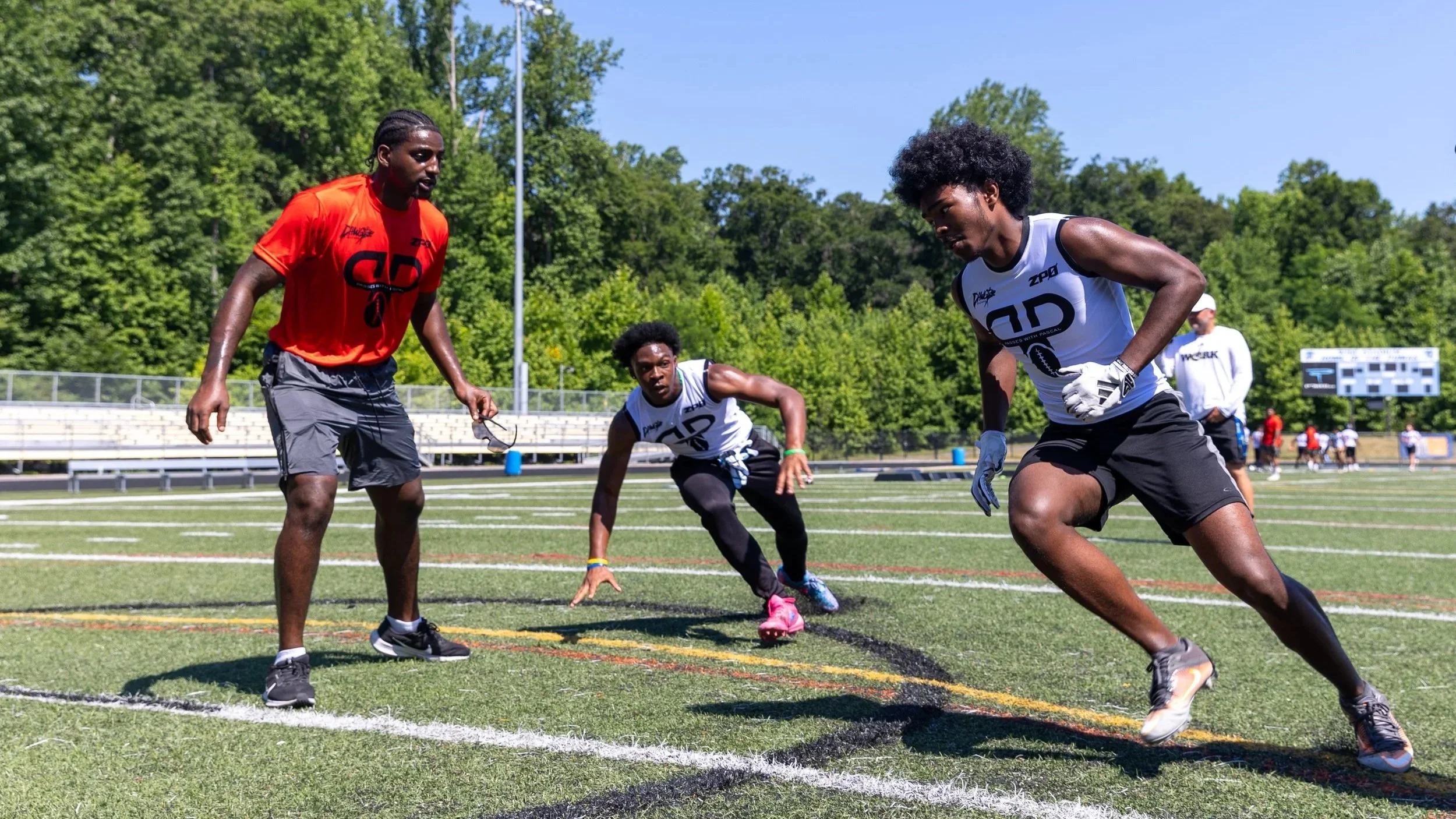 Young male athletes participating in a track and field sprinting event on a green field on a sunny day, with trees in the background.