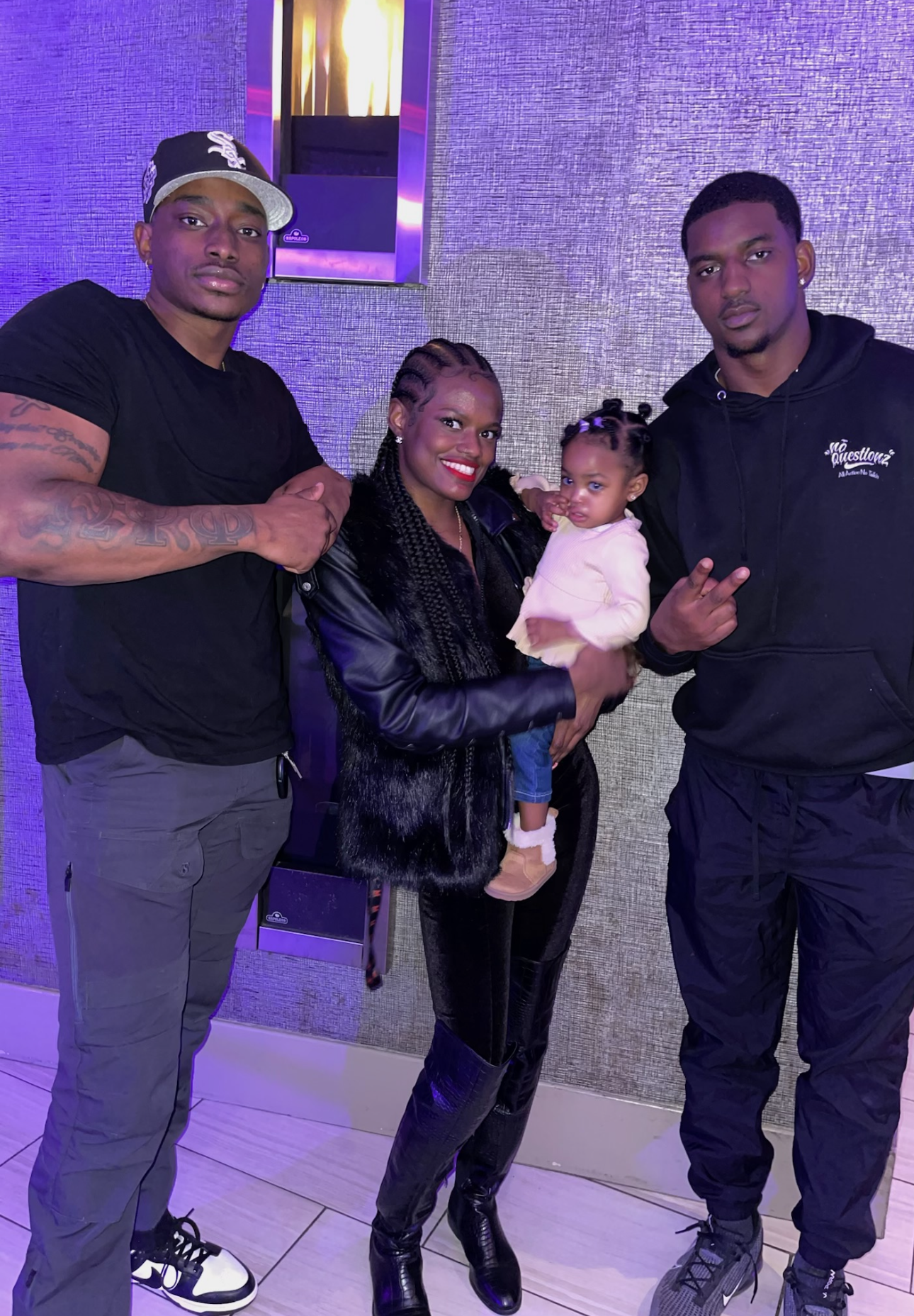 A group of four people, three adults and one child, posing together indoors. The woman in the middle is holding the child, and the man on the right is making a peace sign with his hand. The background features a textured wall and metal paneling.
