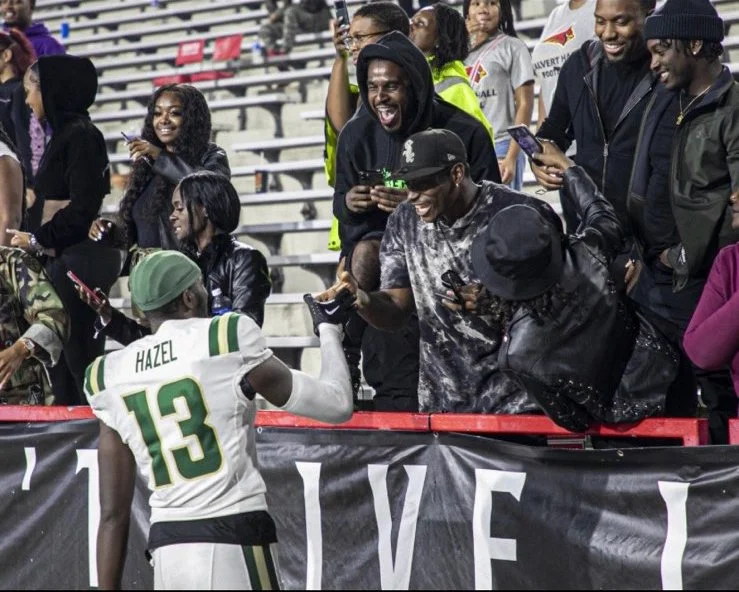 Izaiah Hazel, football player wearing jersey number 13, high-fiving fans in the stands after a game. Fans are smiling, holding phones, and reaching out to shake hands.