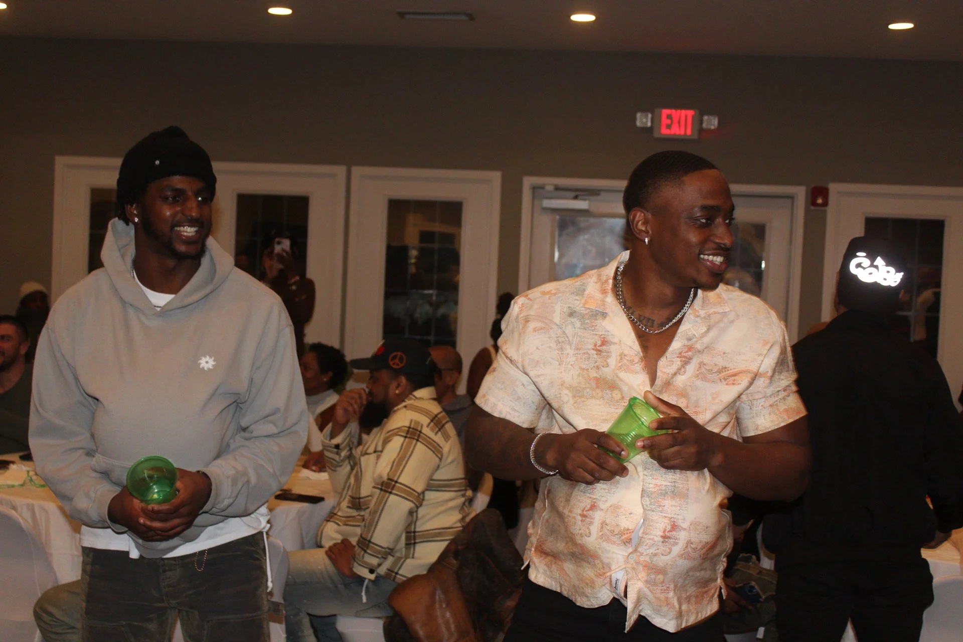 Two young men standing indoors, holding green cups, smiling, with other people sitting at tables in the background.