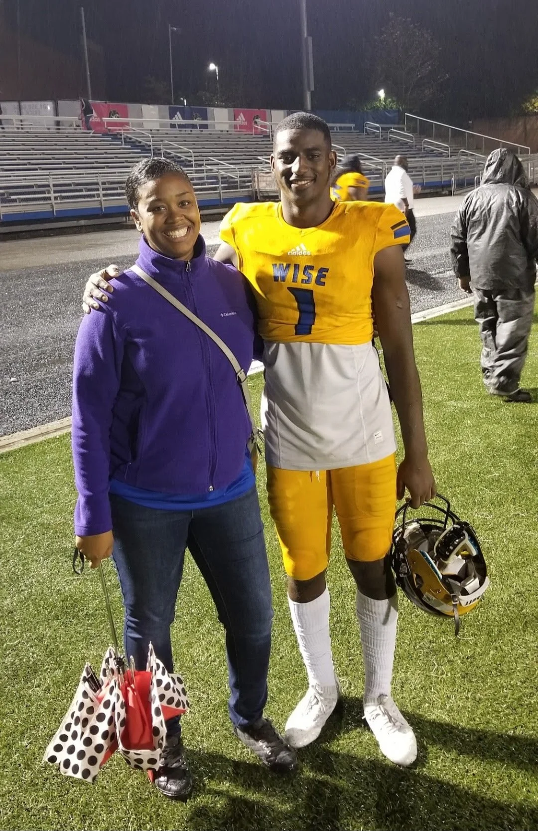 A smiling woman in a purple jacket and jeans standing next to a male football player in a yellow uniform with the number 1, holding a helmet, on a football field at night.