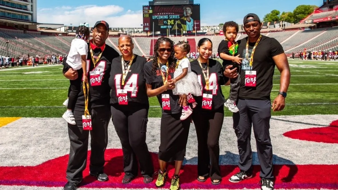 Group of five adults and two children standing on a football field in a stadium, wearing Black and Red NFL jerseys, smiling for a photo.