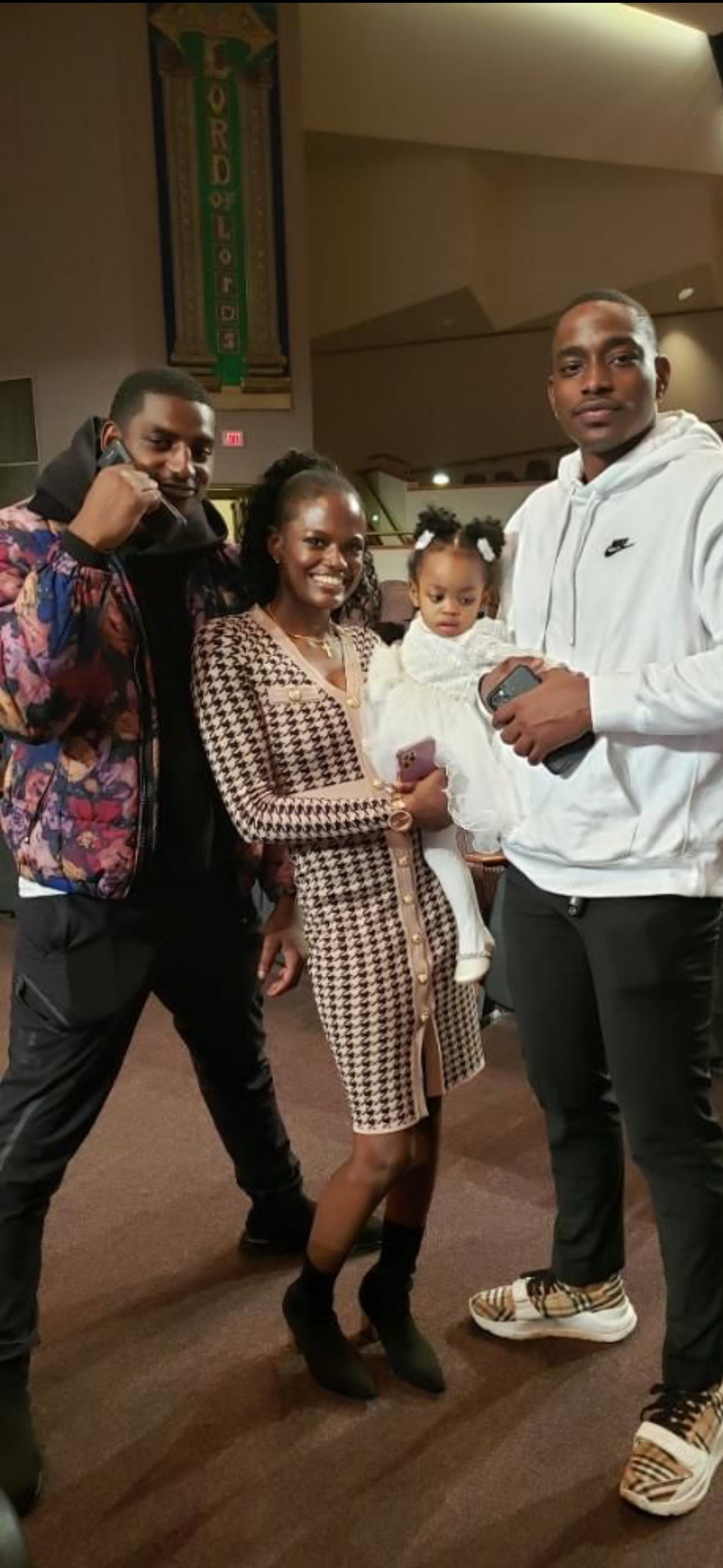 Group of five people, including two men, one woman, and a young girl, posing together indoors. One man is holding a phone to his ear, and the other is holding a phone. The woman is smiling, and the young girl is wearing a white dress with puffs in he
