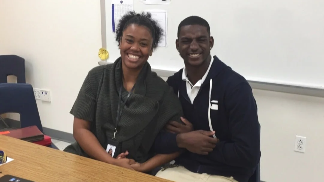 Two smiling young adults sitting at a table in a classroom with a whiteboard behind them.