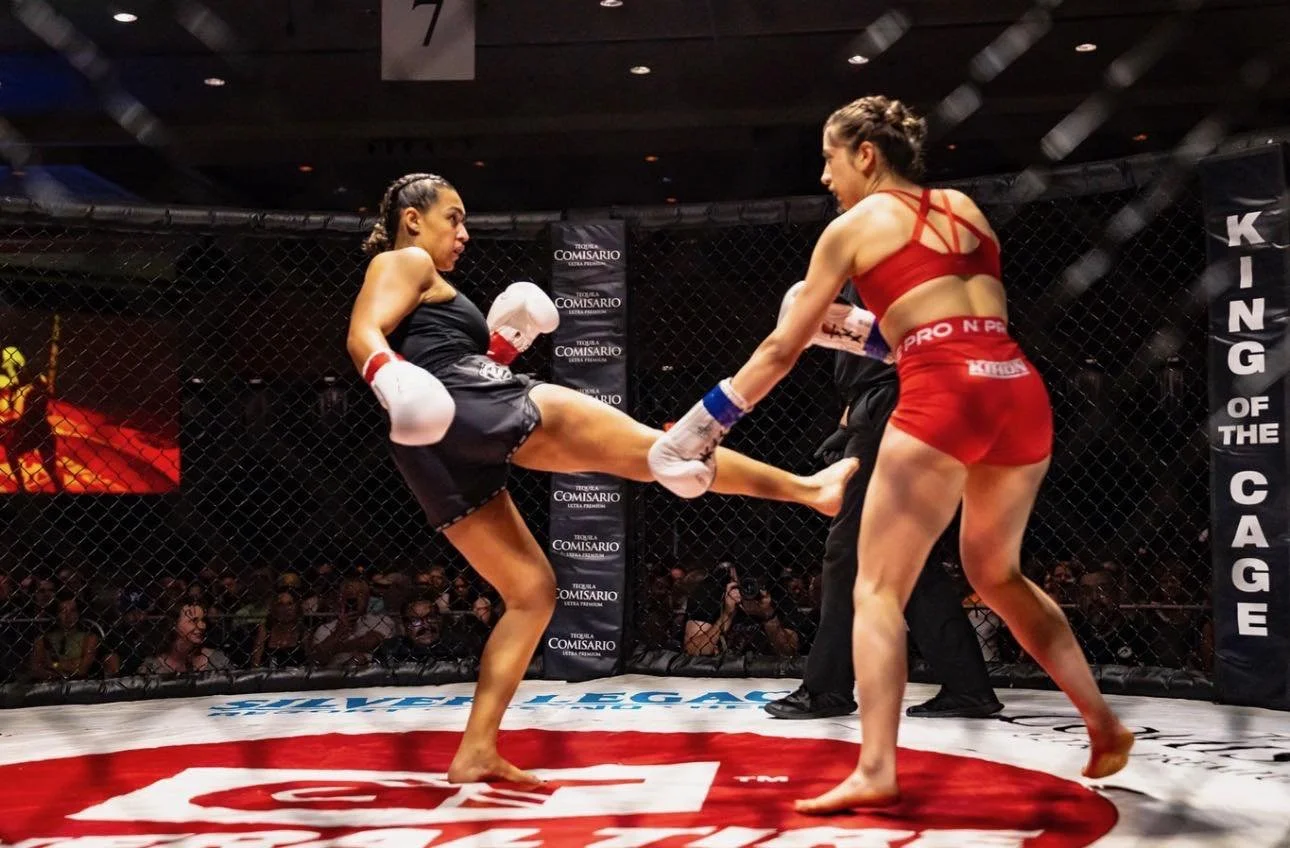 Two women fighters in a mixed martial arts fight inside a cage, with one woman delivering a high kick to the other's midsection, while a referee observes. The audience is visible in the background.