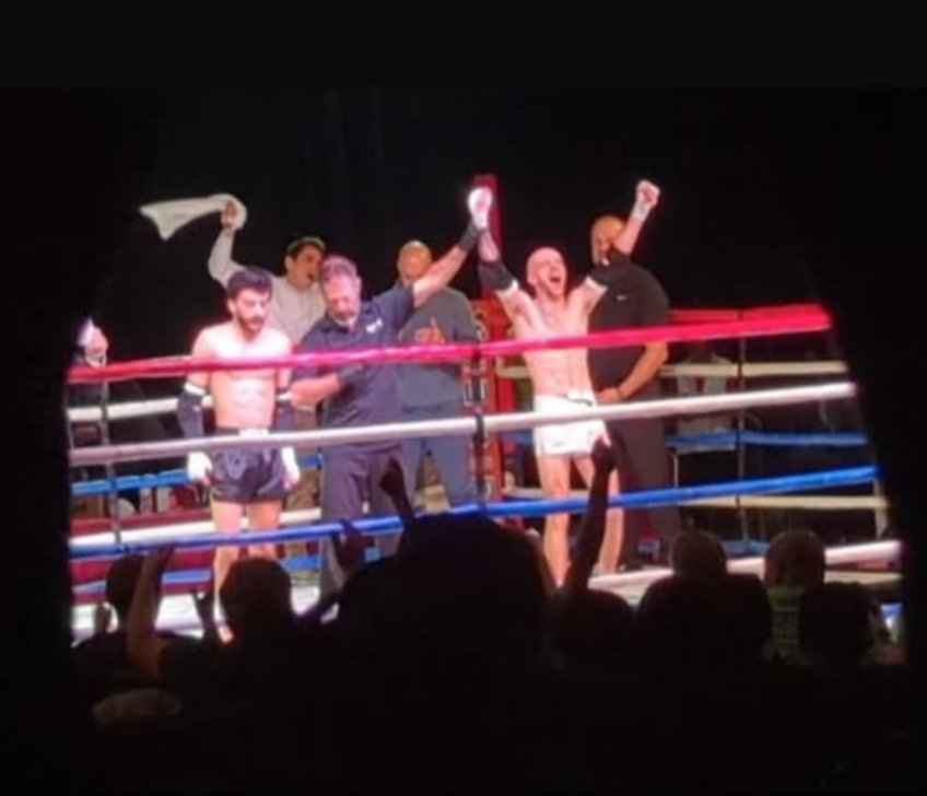 Boxers and officials celebrate in a boxing ring after a match, with audience members visible in the foreground.