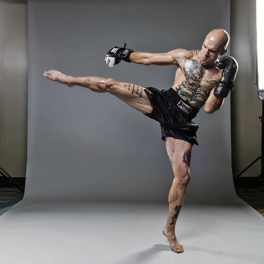 Zach Bunnell demonstrating a muay thai kick , wearing black boxing shorts and black boxing gloves.