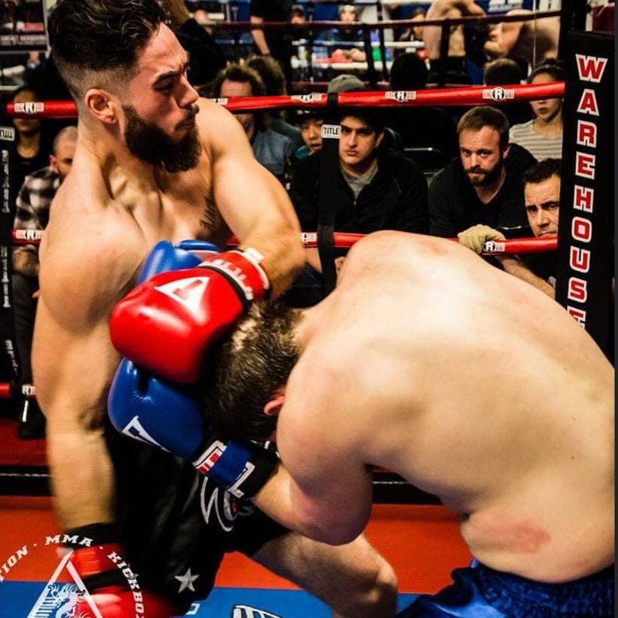 Two shirtless male fighters in a boxing or MMA ring engaged in a fight, with one delivering a punch to the other's head. The fighter throwing the punch has a beard and is wearing red and blue gloves. The opponent's head is bent downward, and he is we