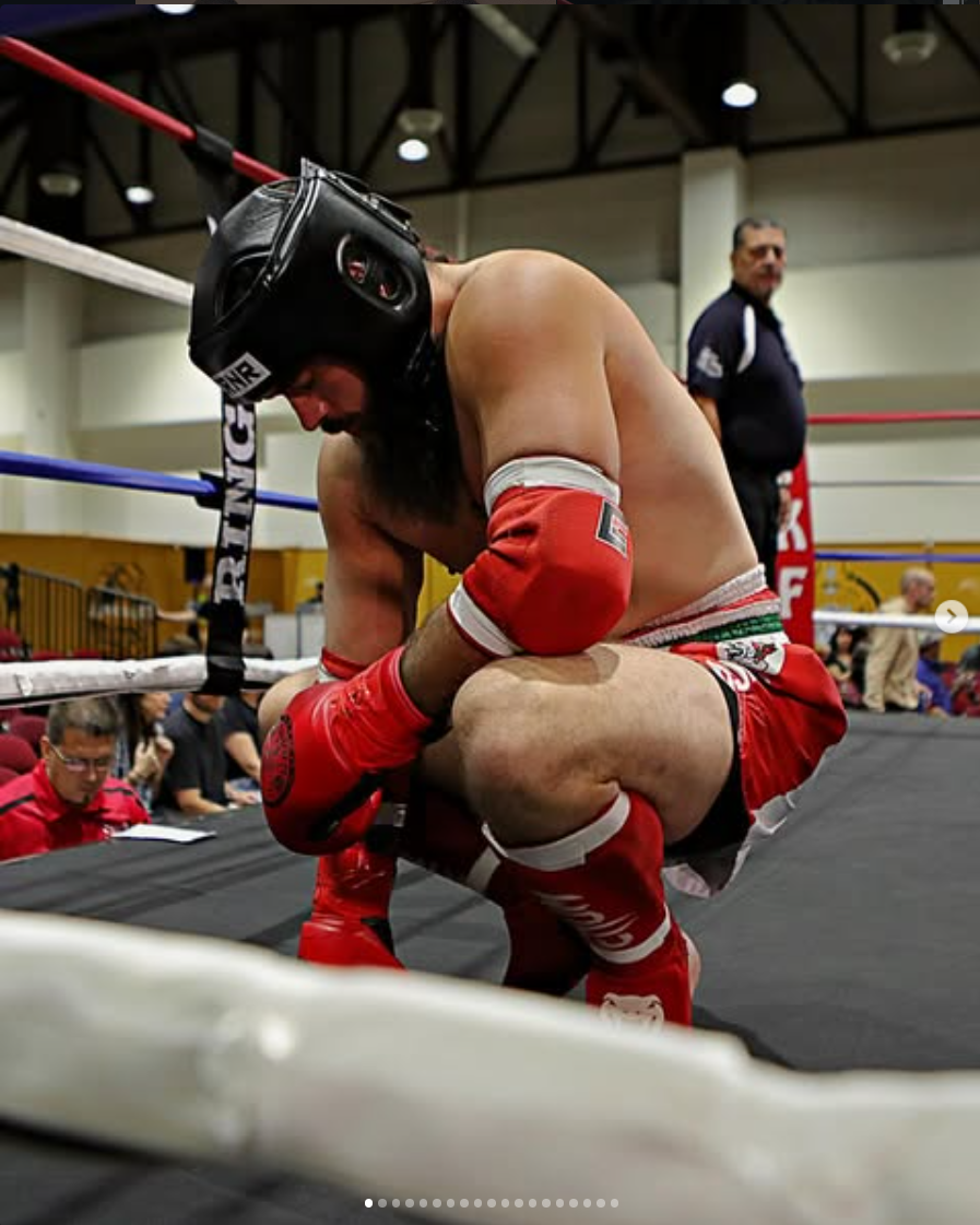 A male boxer kneeling in the corner of a boxing ring, wearing red boxing gloves, red shorts, and a black headgear. He appears to be resting or praying. In the background, a man in a dark shirt stands, and there are people sitting ringside.