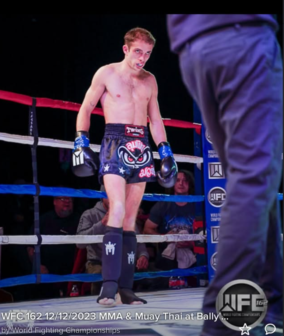 A shirtless male Muay Thai fighter standing in a boxing ring, wearing black shorts and gloves, with his head tilted down and a intense expression. The ring has red, white, and blue ropes. Spectators are visible in the background.
