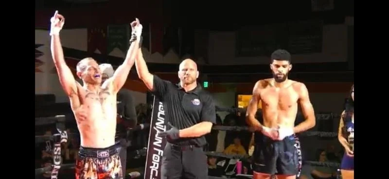 A victorious Muay Thai fighter with tattoos and a headband celebrating with raised arms, a referee holding his hand, and another fighter standing nearby in a boxing ring.