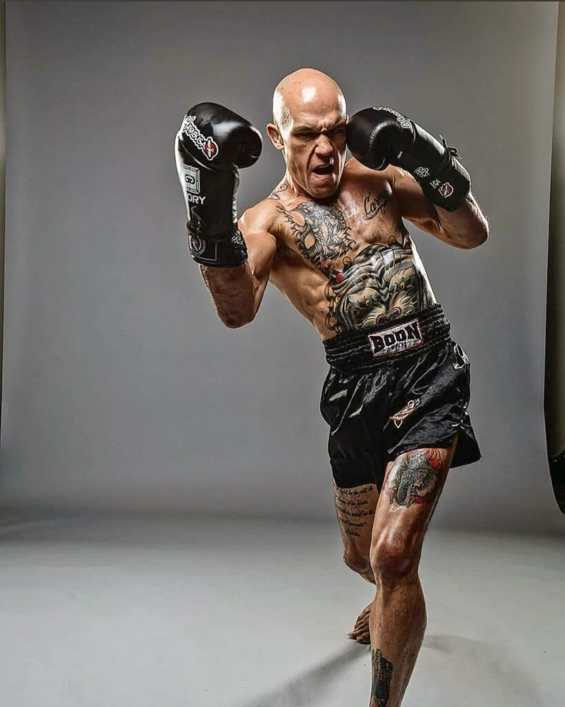 A tattooed male boxer wearing black gloves and shorts, posing in a fighting stance against a plain gray background.
