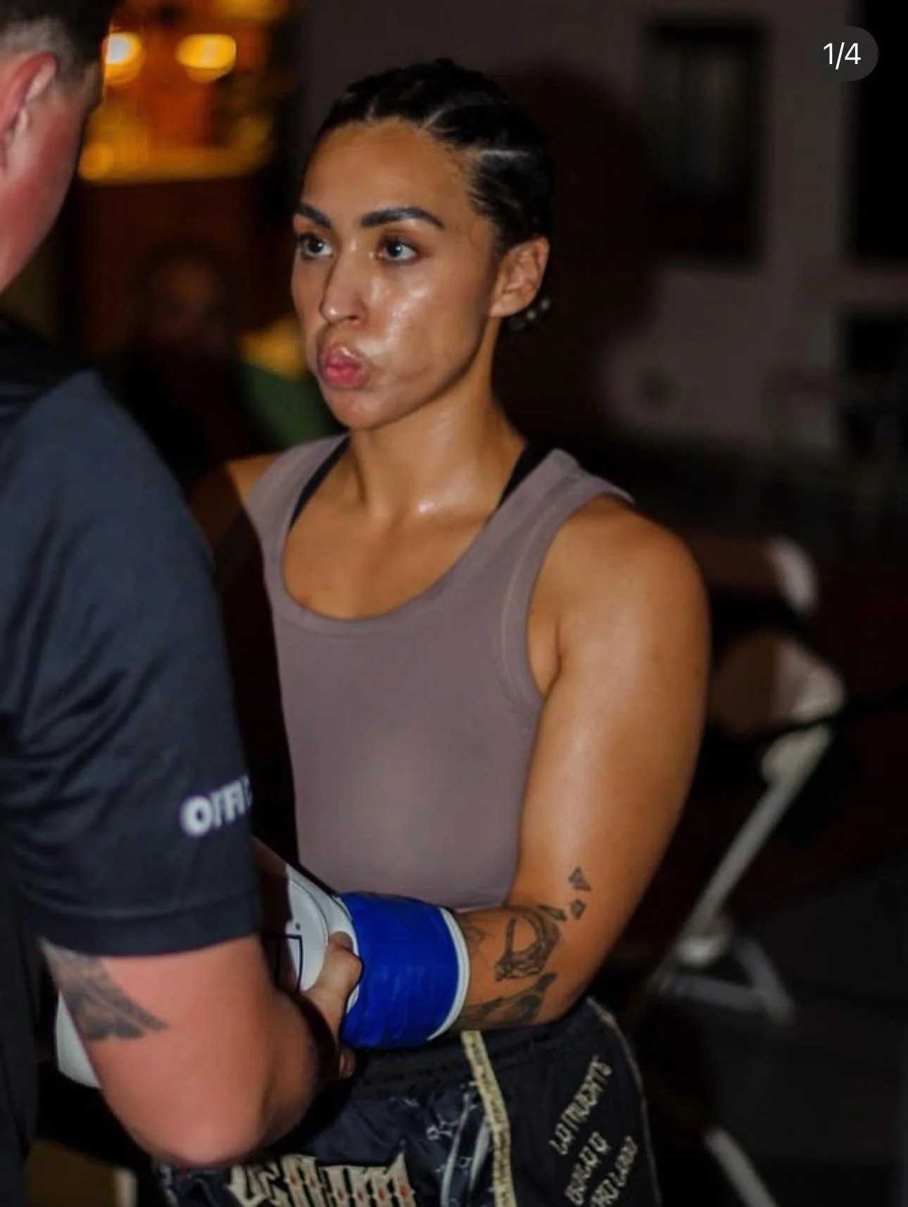 A woman with tattoos on her left arm and slicked-back hair, wearing a beige tank top, standing in an indoor gym.