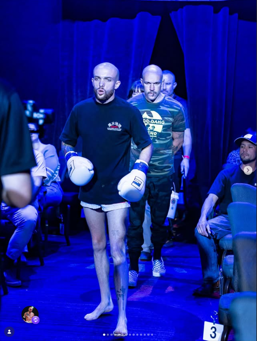 A boxer with tattoos and boxing gloves walking through a blue-lit room, followed by a man in a camouflage shirt and other spectators seated nearby.