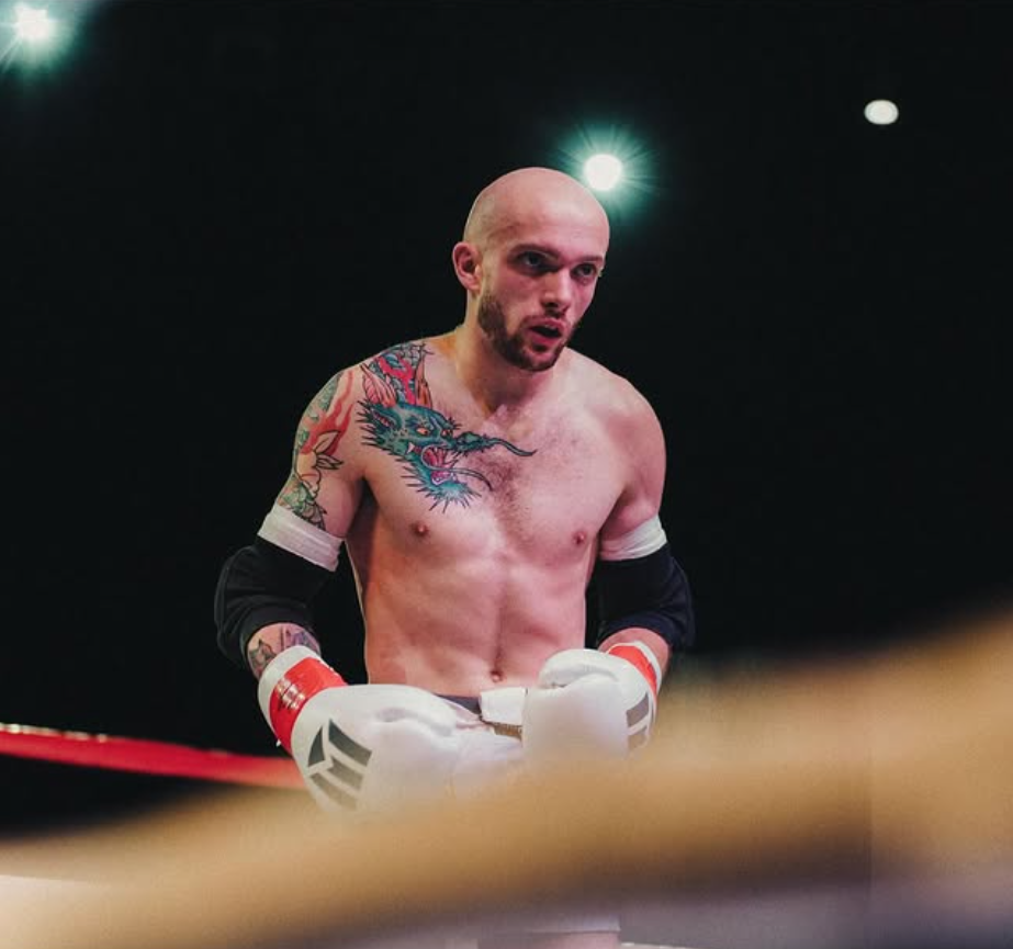 Colin, wearing boxing gloves and protective arm gear, stands inside a boxing ring with a dark background and bright overhead lights.
