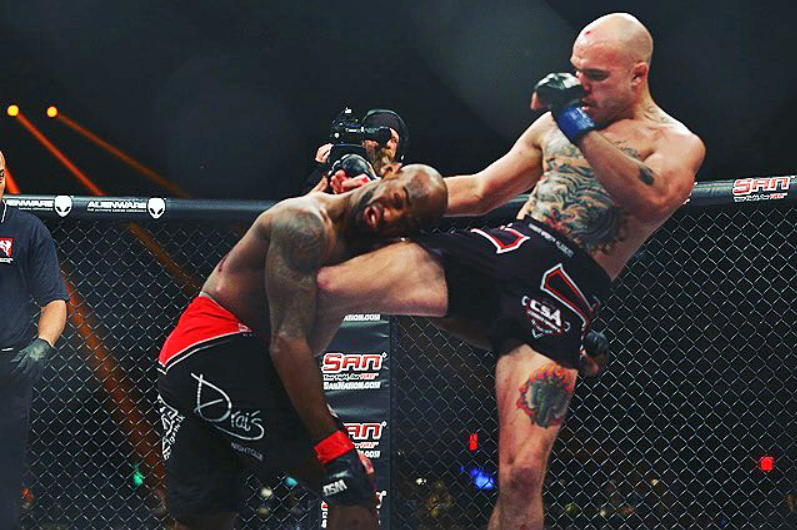Mixed martial arts fighters in a cage during a match, one fighter delivering a high kick to the other's head.