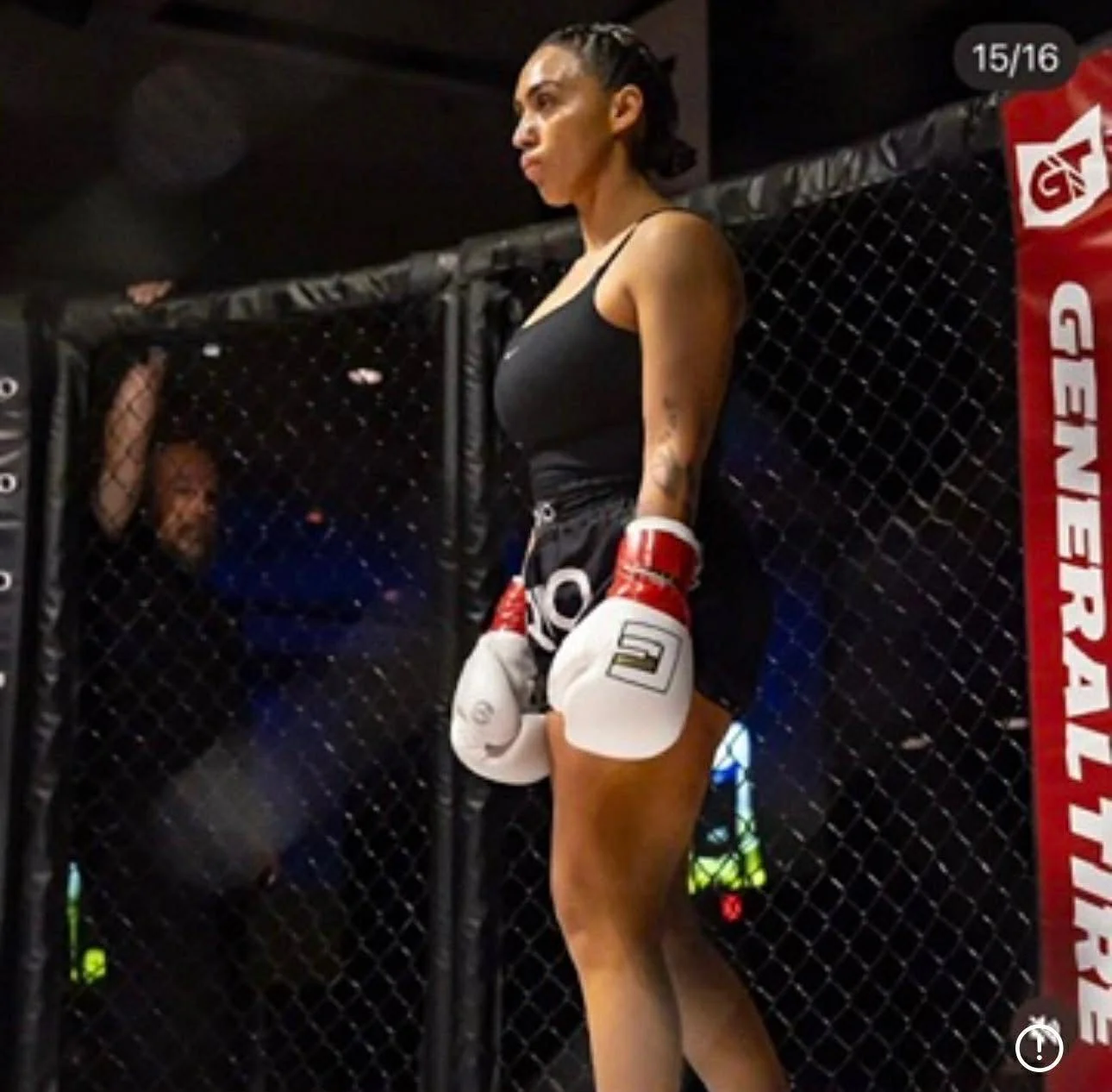 Female mixed martial artist standing against a cage fence, wearing black sportswear and white MMA gloves, inside a UFC octagon with a referee watching in the background.