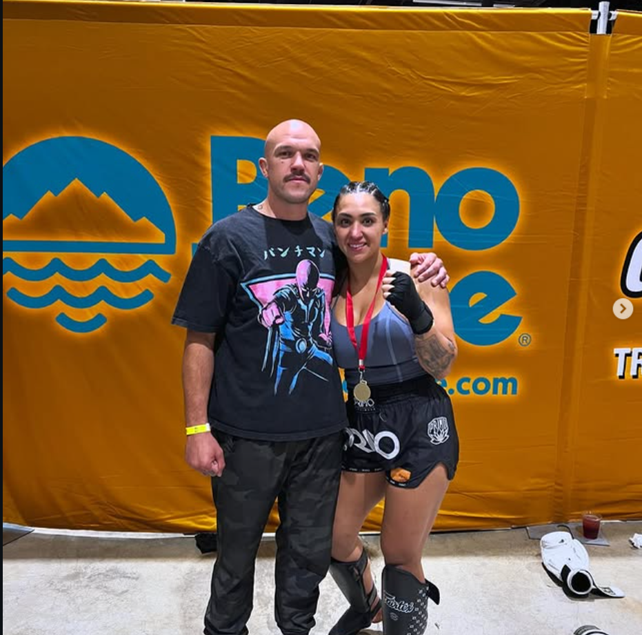 A man and a woman posing together after a martial arts or boxing event, with a large yellow banner in the background. The woman is wearing fight shorts, a top, ankle support, and has a medal around her neck, while the man is wearing casual clothing a