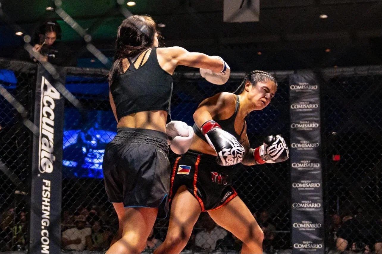 Two women fighters in a mixed martial arts cage during a fight. One is throwing a punch, the other is reacting defensively. Both wear black athletic clothing and gloves. The cage has a black chain-link fence with advertisements.