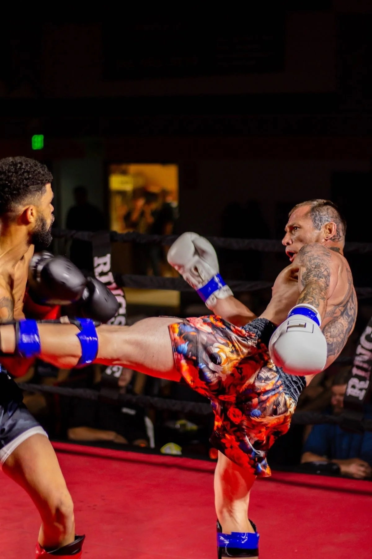 Two men engaged in a Muay Thai fighting match in a boxing ring. The man on the right is delivering a high kick to the man on the left. Both are wearing boxing gloves and athletic shorts, with the man on the right having tattoos on his arm and chest.