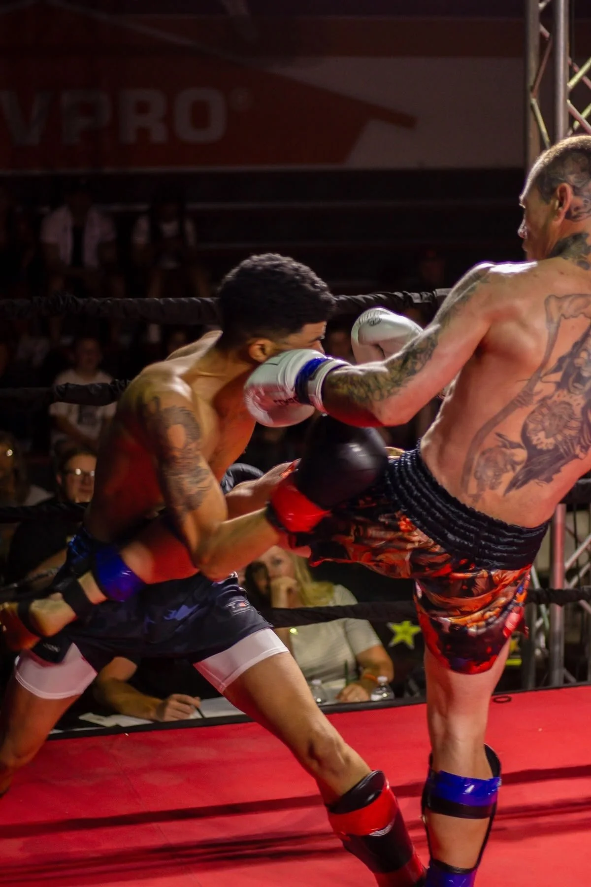 Two shirtless male fighters with tattoos competing in a boxing match inside a ring. One fighter is delivering a high kick to the other's face, while the opponent braces for impact.