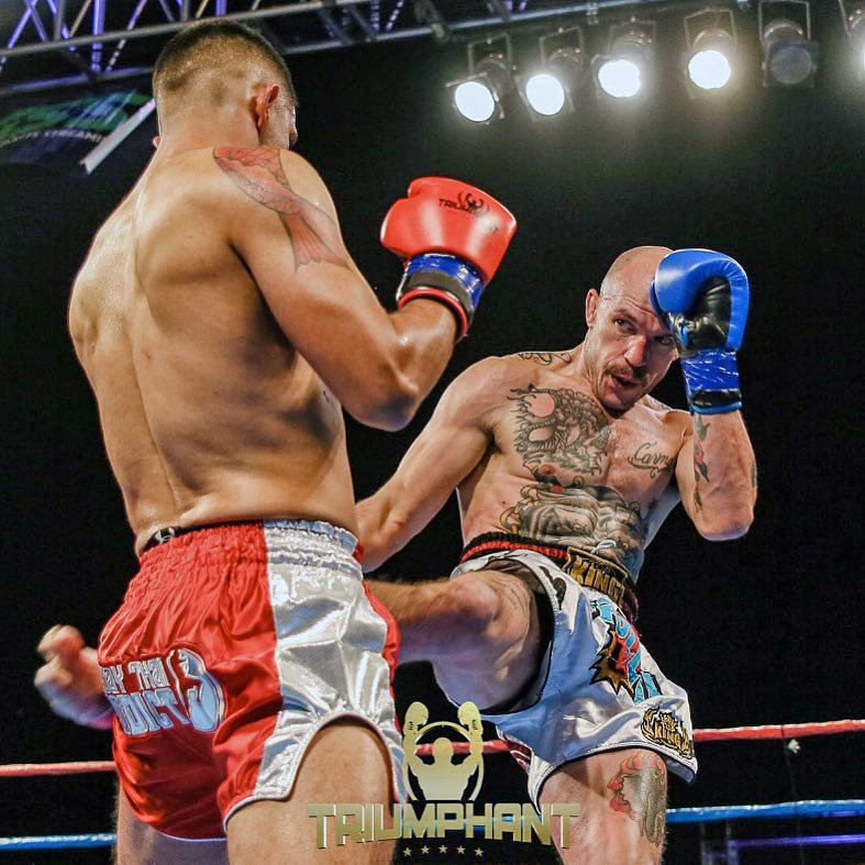 Two male fighters in a boxing ring engaged in a match, one with red gloves and the other with blue gloves. The fighter with blue gloves is delivering a kick to the midsection of the fighter with red gloves. Bright overhead lights illuminate the scene.