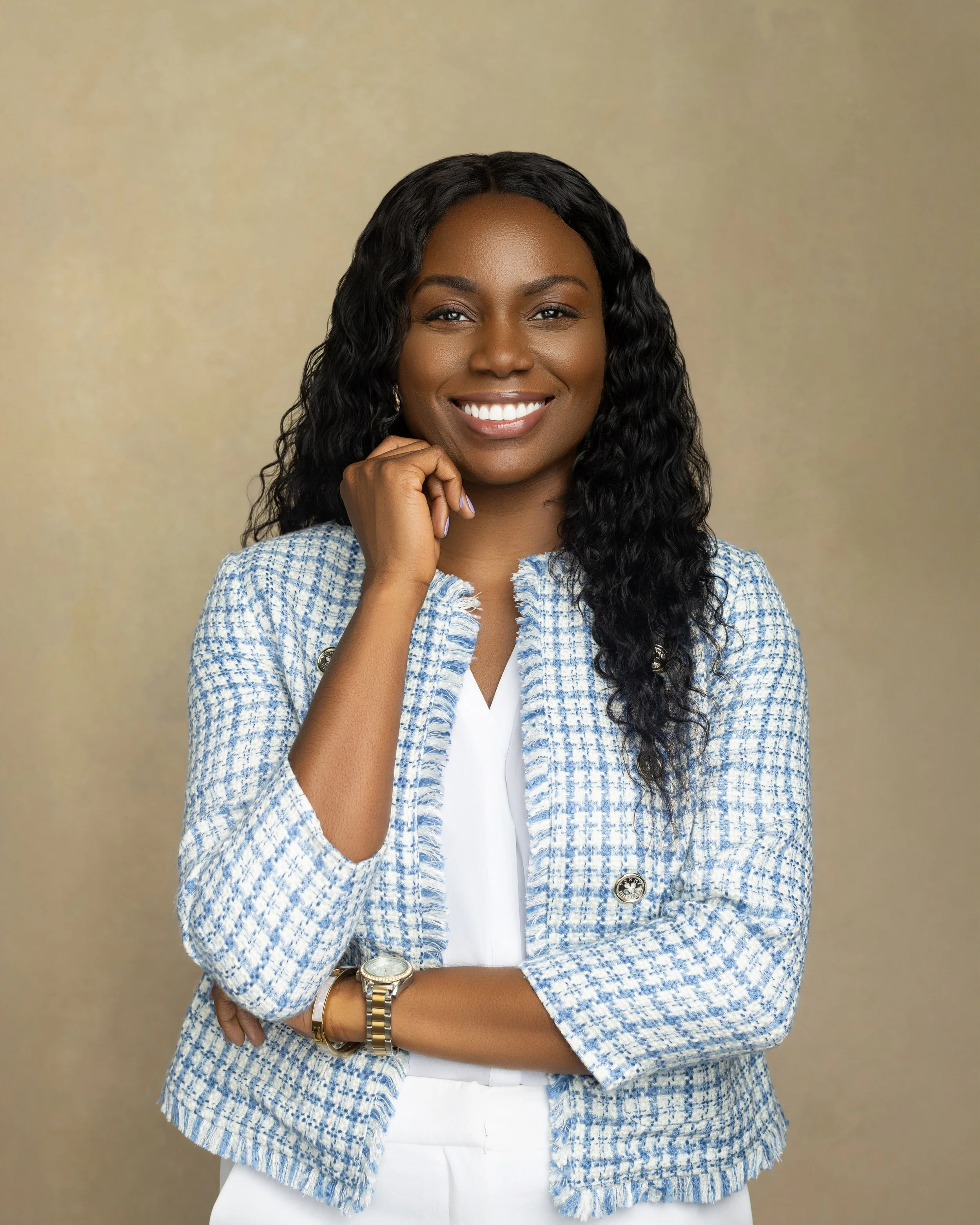A smiling woman with long curly black hair, wearing a blue and white tweed blazer, white blouse, white pants, a gold wristwatch, and earrings, standing against a beige background.