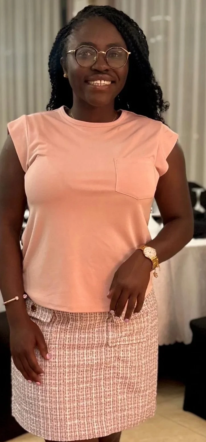 A young woman with glasses, wearing a pink sleeveless top and a patterned skirt, standing indoors with a smile.