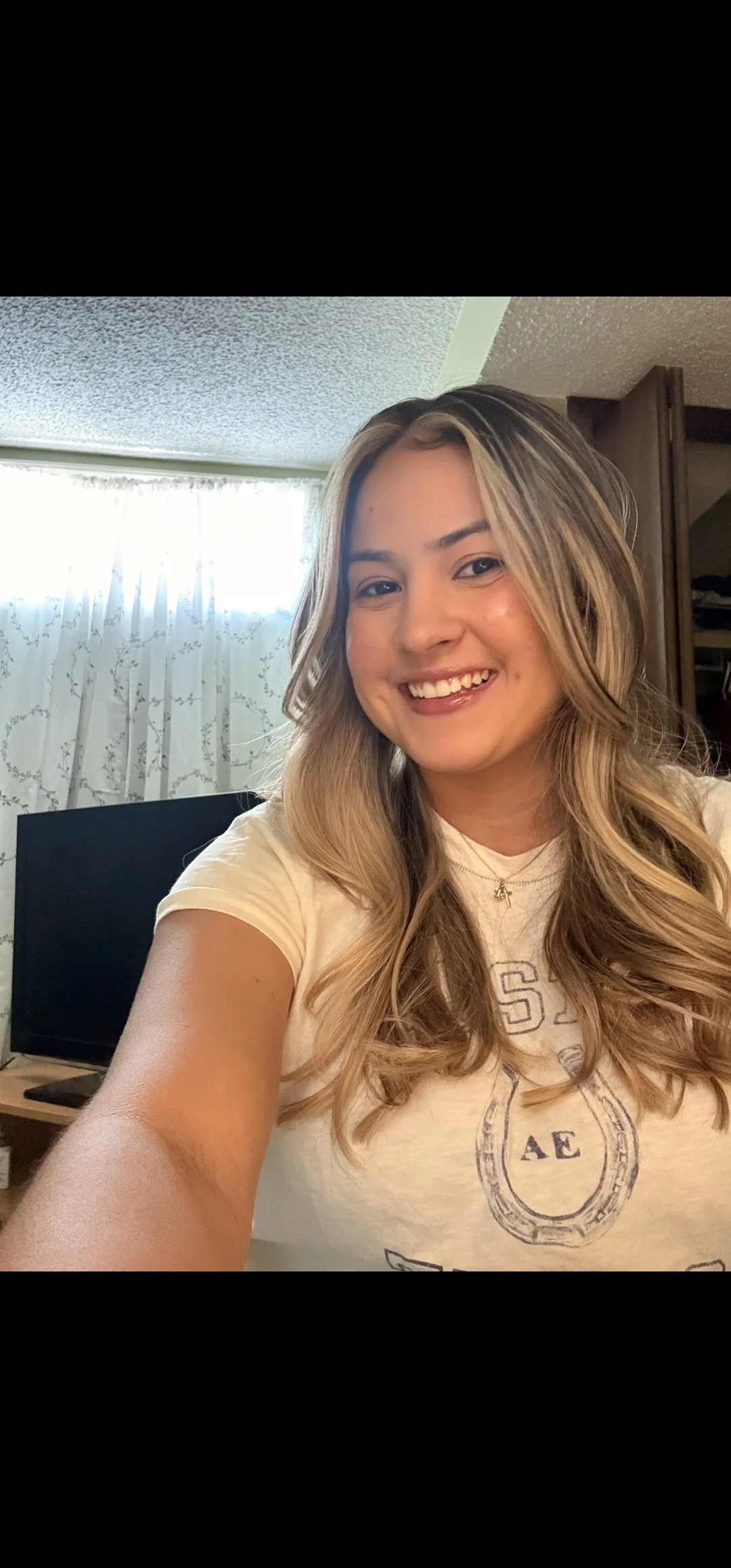 A young woman with long, wavy blonde hair smiling in a room with a window with sheer curtains and a flat-screen TV in the background.
