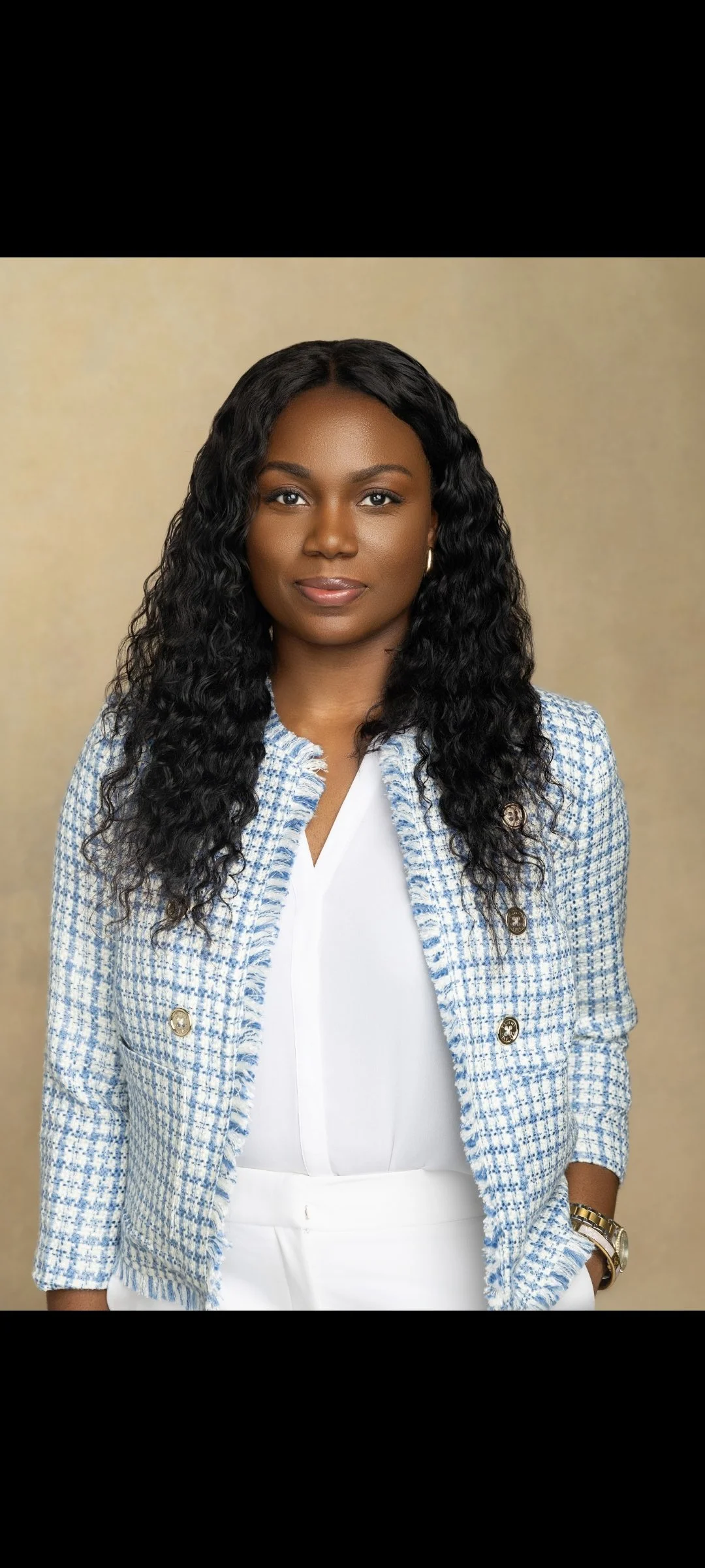 A woman with dark, curly hair wearing a light blue and white checked blazer, white top, and white pants, with a gold watch and earrings, standing against a neutral background.