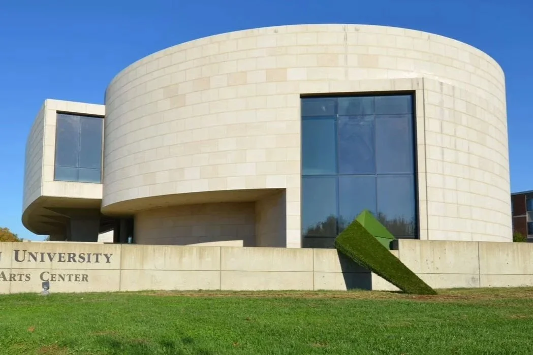 Modern university arts center with curved concrete facade and large glass window, featuring a neatly trimmed pyramid-shaped hedge in front.