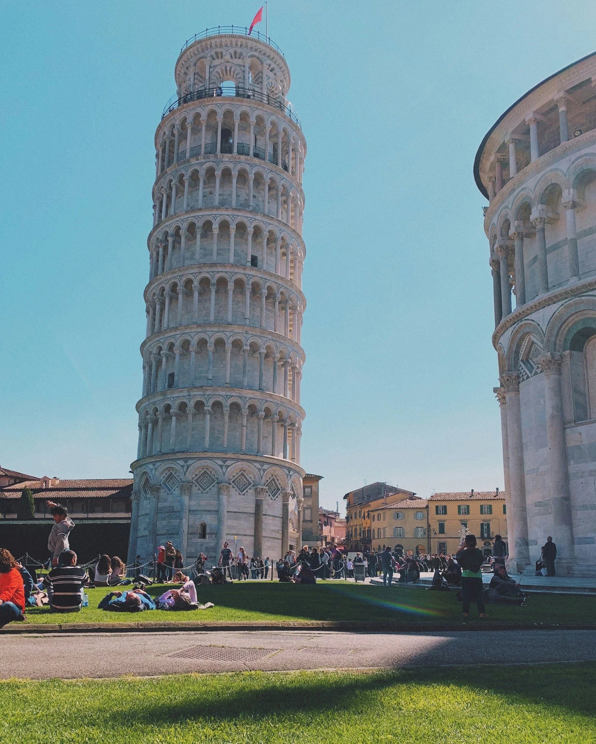 People sitting on a lawn in front of the Leaning Tower of Pisa, Italy, with a clear blue sky in the background.