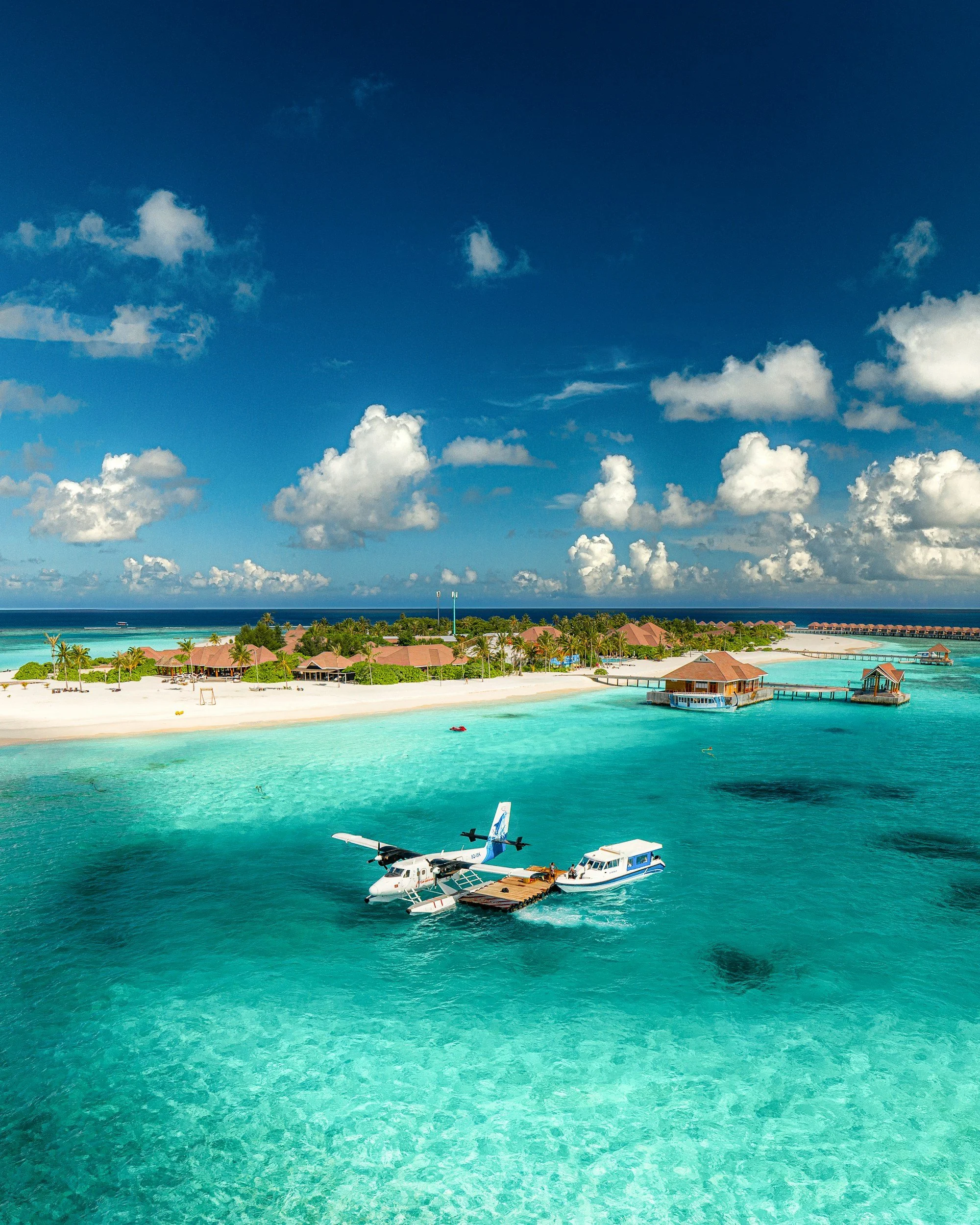 Tropical beach with turquoise water, overwater bungalows, and a seaplane flying low over the water under a blue sky with scattered clouds.