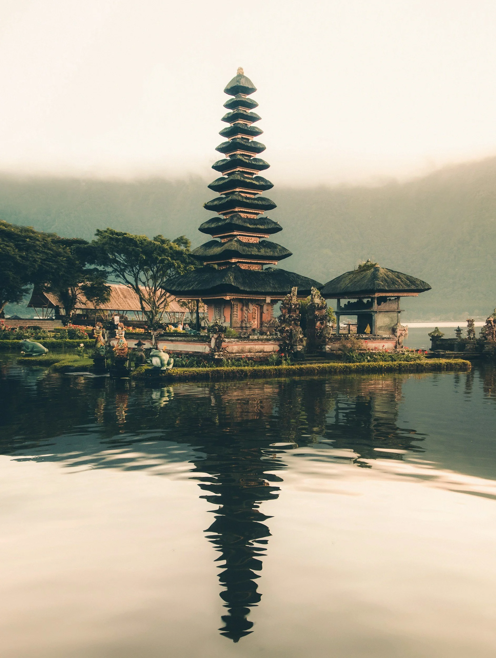 A traditional Balinese multi-tiered pagoda with a thatched roof, situated on the edge of a body of water, with lush green hills and mist in the background.