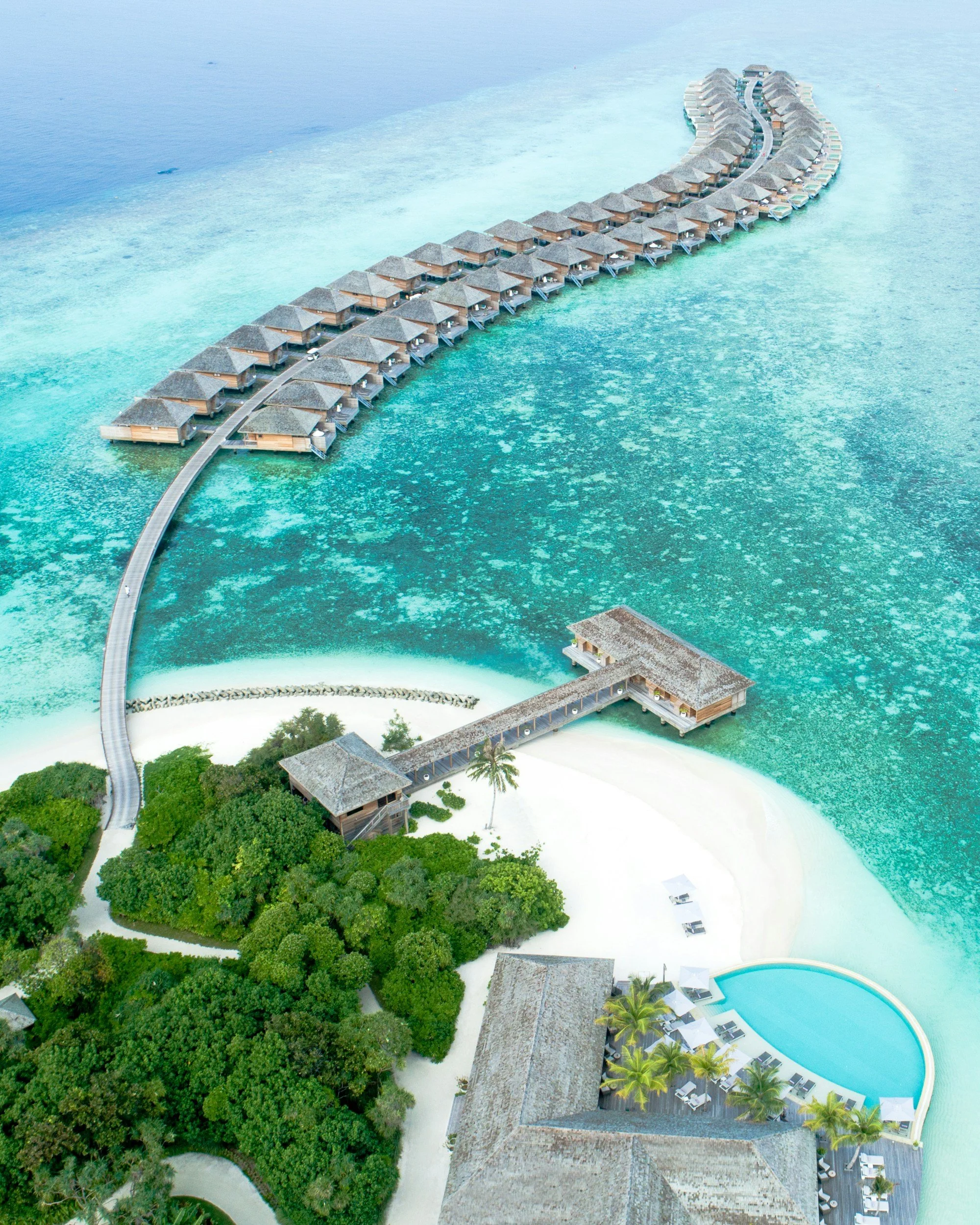 Aerial view of a resort with overwater bungalows, a lagoon, and a swimming pool surrounded by lush greenery and white sandy beaches.