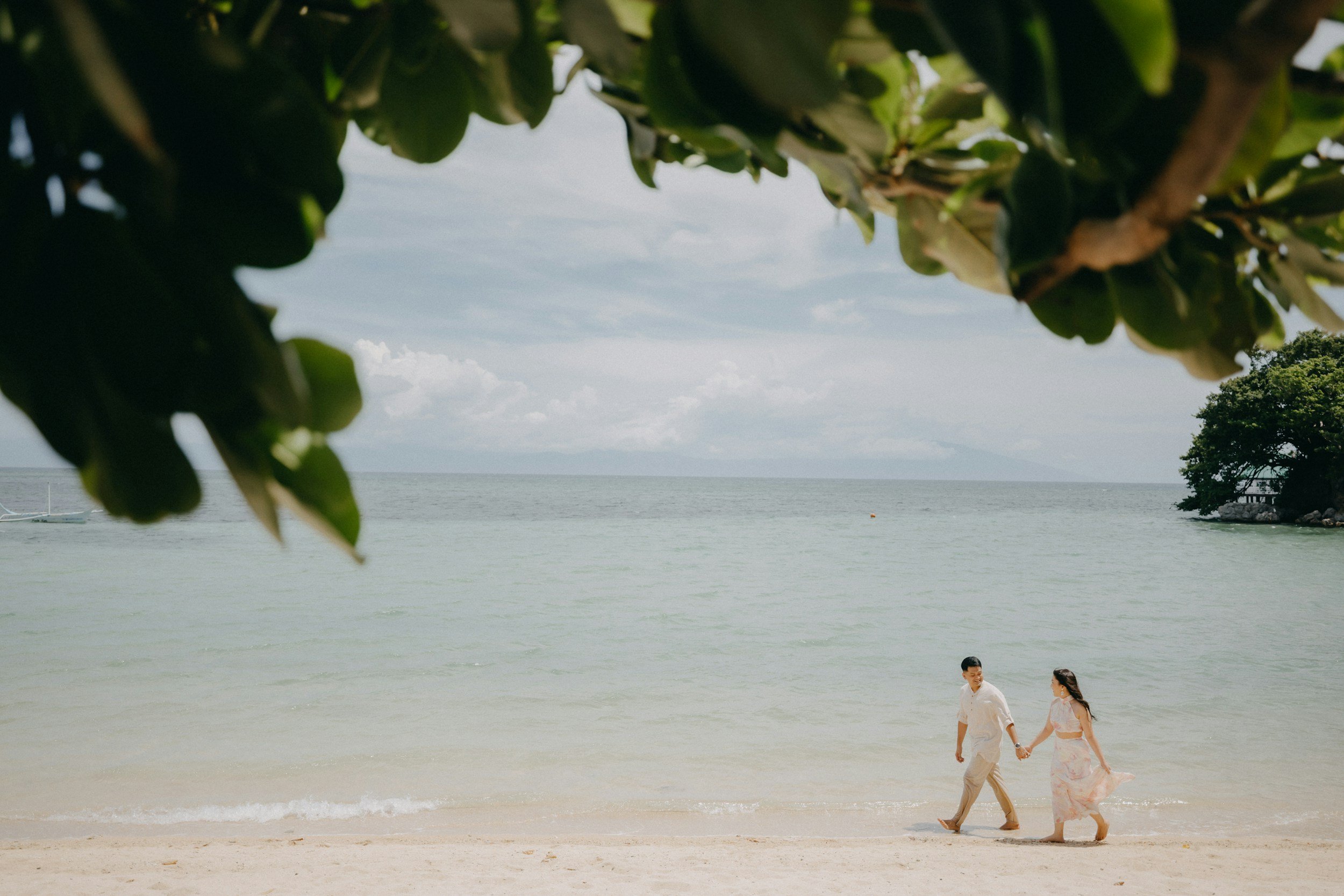A couple walking hand-in-hand along the beach with the ocean in the background and trees framing the top of the image.