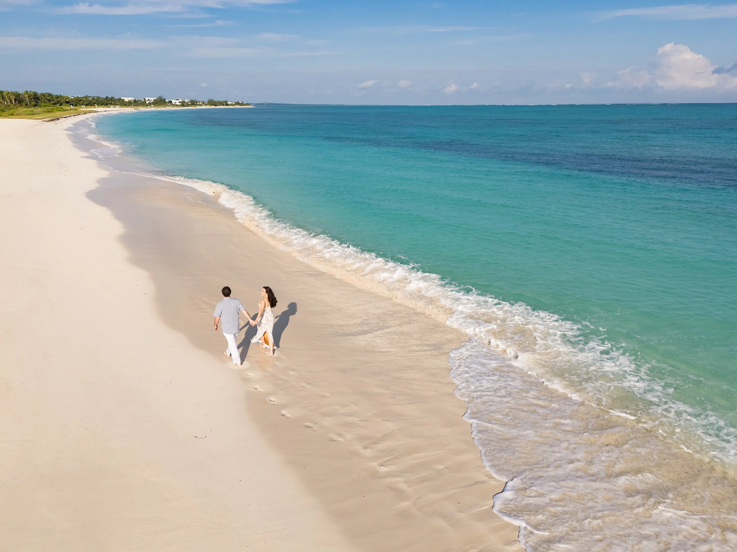 A couple walking along a sandy beach with turquoise water and a distant shoreline with greenery and buildings on a sunny day.