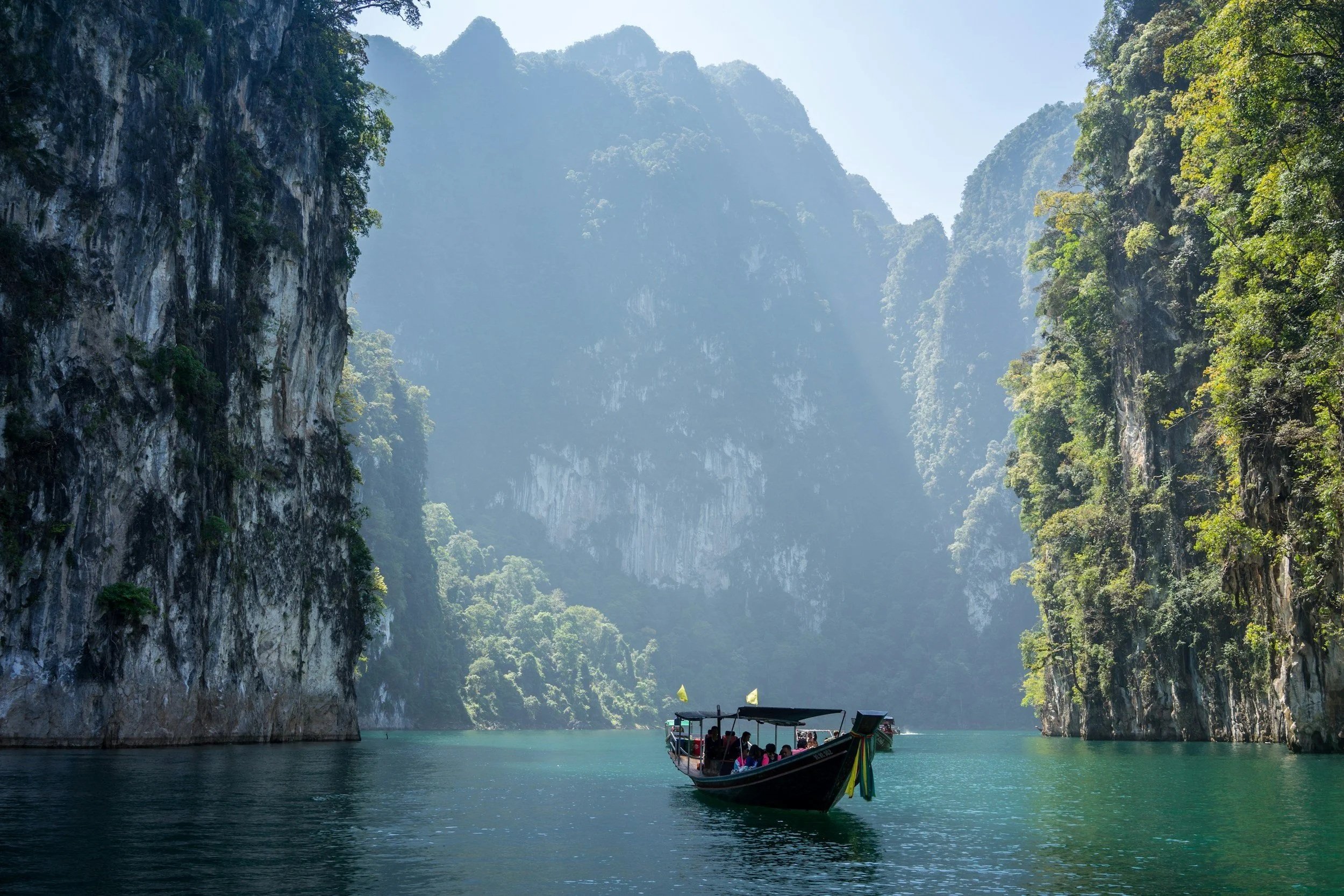 A boat with tourists traveling through a narrow, lush green canyon with tall cliffs on either side and mist in the background.