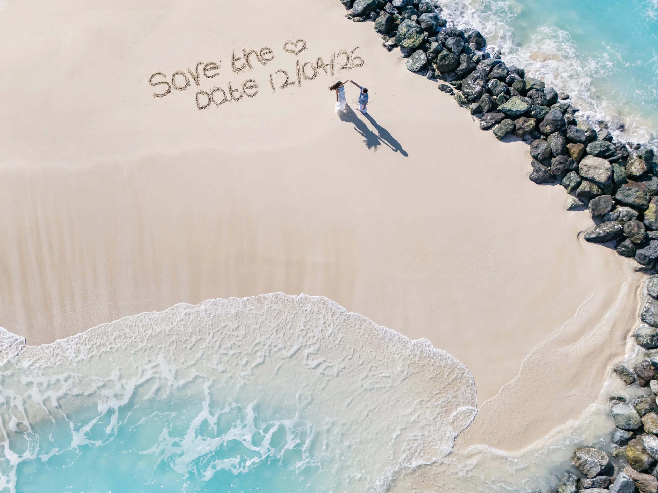 Aerial view of a sandy beach with waves crashing onto shore and a rock jetty along the right side. Sandy writing on the beach says 'Save the ❤️ Date 12/04/26'.