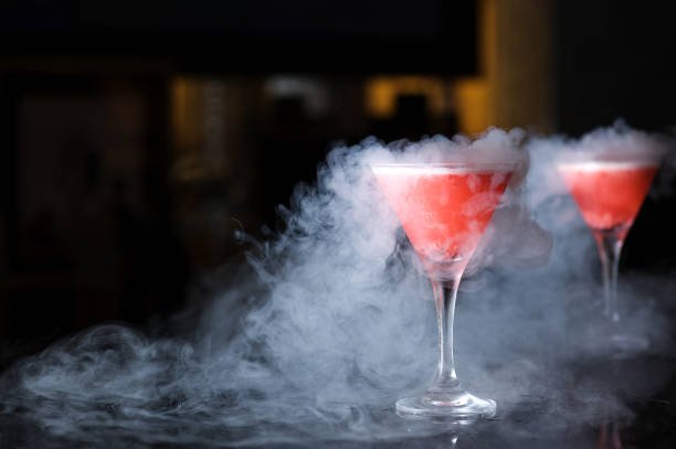 Two pink cocktails in martini glasses with smoke or fog around them on a dark surface.