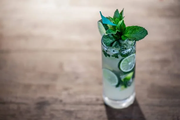A tall glass of mojito cocktail with lime slices, mint leaves, and ice on a wooden surface.