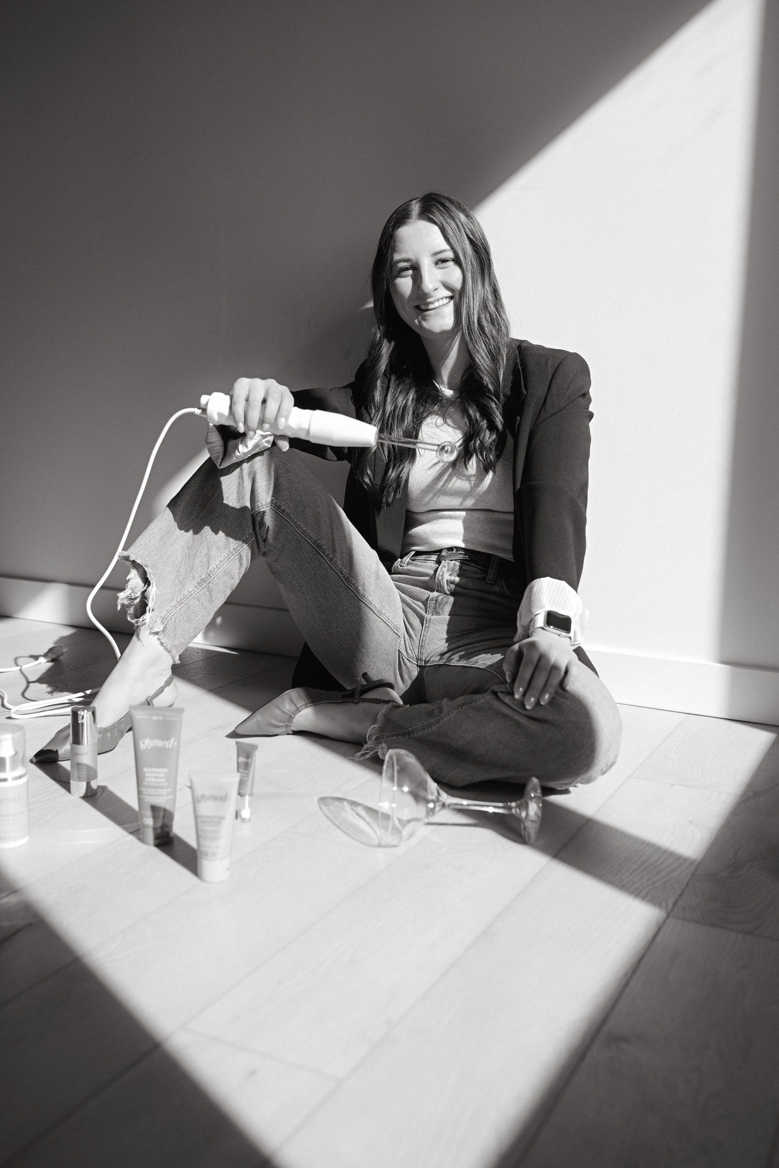 A woman with long dark hair, sitting on the floor, appears to be applying makeup or skincare products with various bottles and tubes nearby. She is smiling and wearing casual clothes and a blazer, with sunlight and shadows creating a high-contrast, black-and-white scene.