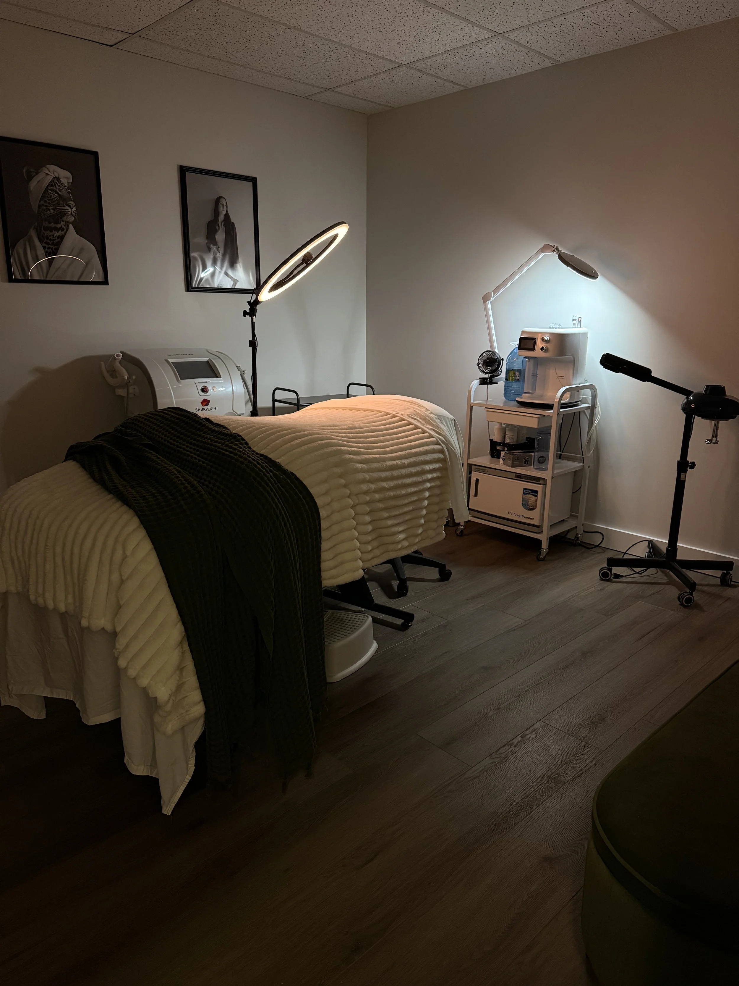 A medical or spa treatment room with a massage or therapy bed covered with a yellow quilt and a dark blanket. To the right, there is medical equipment including a water dispenser, a small fan, and a lamp. On the wall, there are two framed black and white photographs, one of a woman with a leopard print face and the other of a woman in a black jacket. The room has a wood floor and a suspended ceiling.