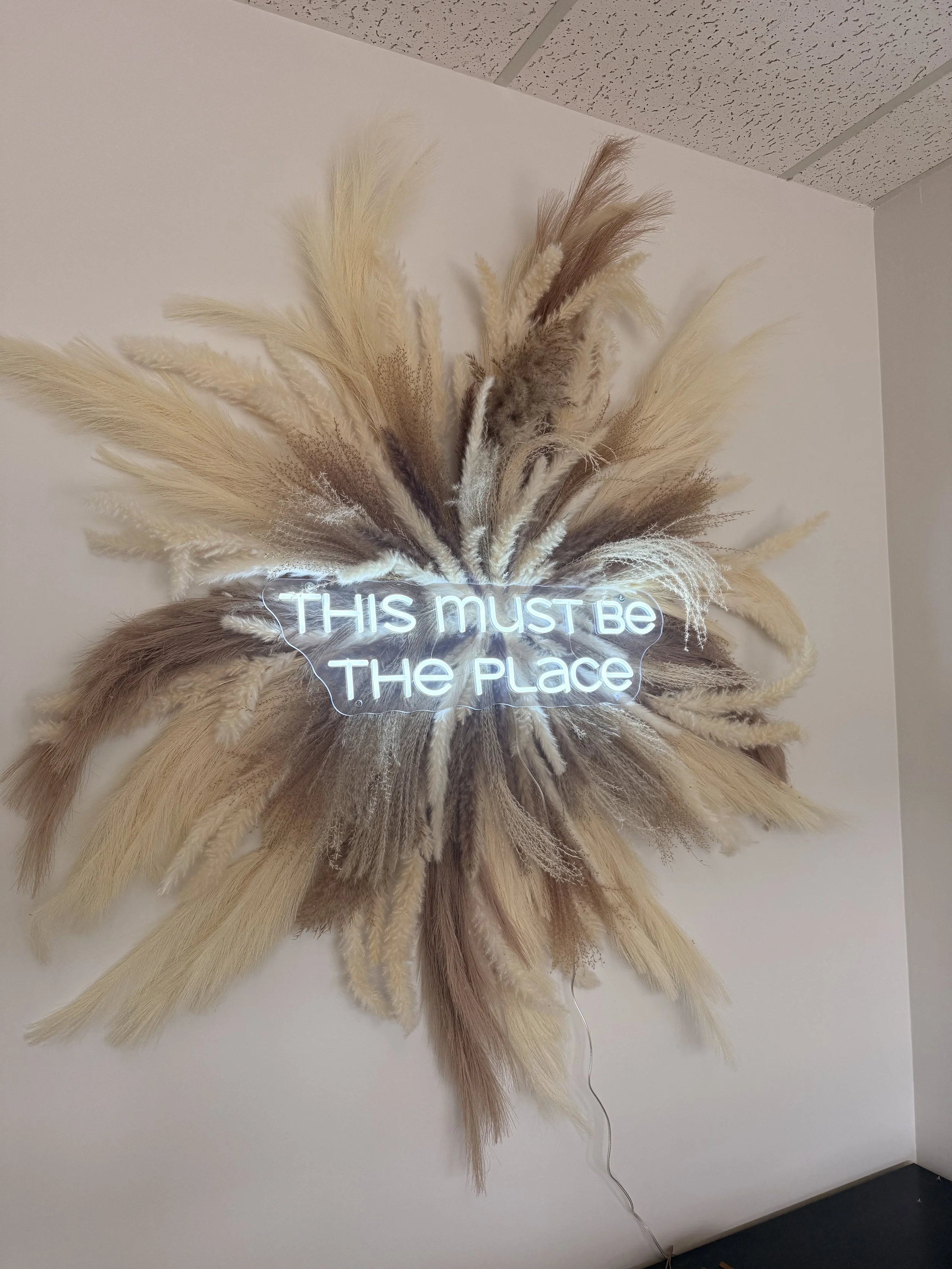 A decorative wall hanging made of dried pampas grass and other dried plants, arranged around a neon sign that reads 'THIS MUST BE THE PLACE,' mounted on a plain wall.