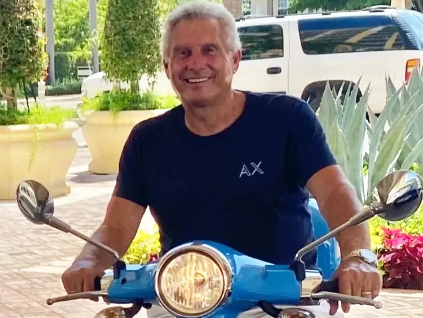 A smiling man with white hair sitting on a blue scooter outdoors surrounded by green plants and flowers, with parked cars in the background.