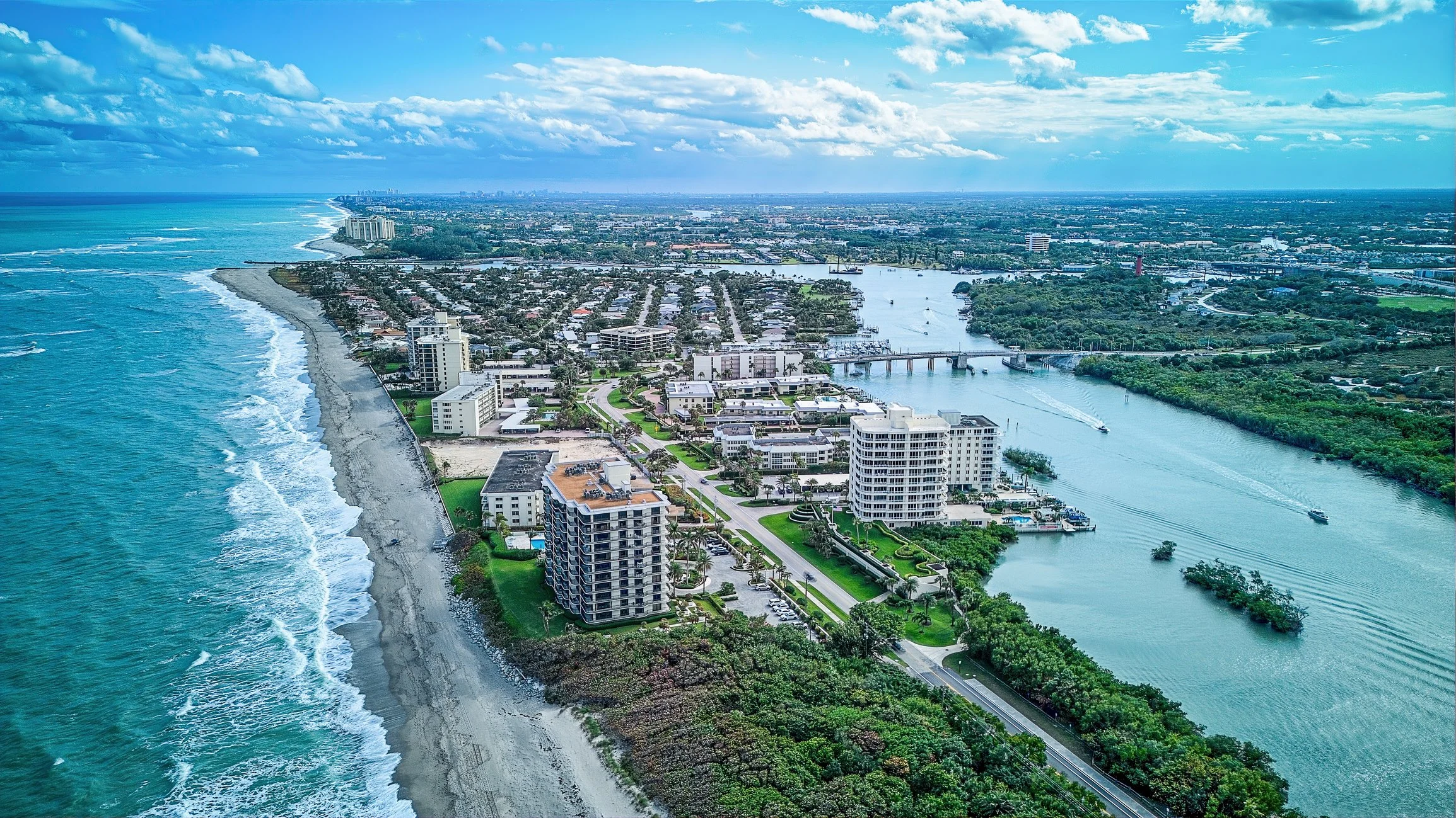 Aerial view of a coastal city with high-rise buildings, residential areas, a river, and the ocean on the left side under partly cloudy skies.