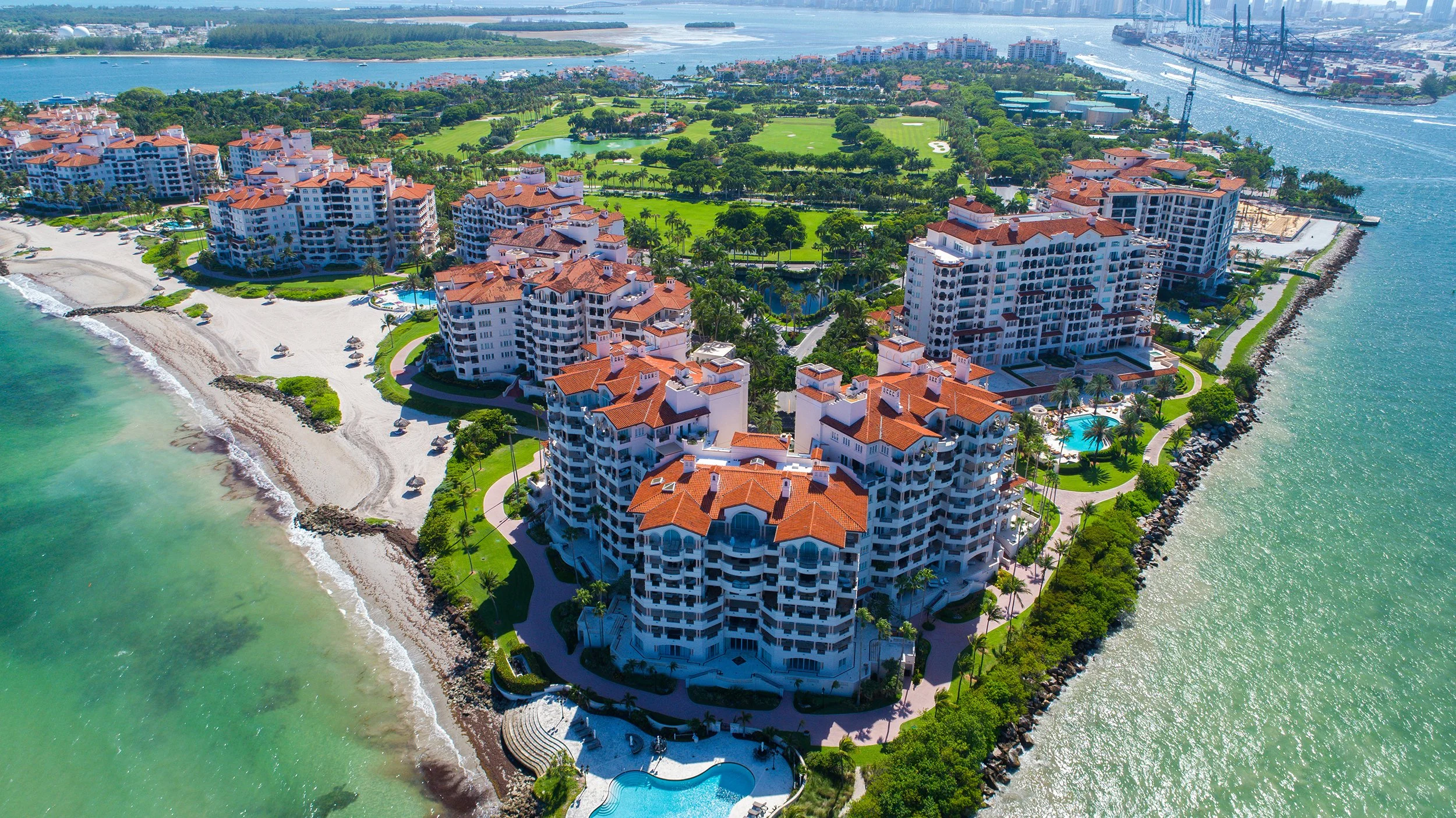Aerial view of a luxurious beachfront resort with multiple white buildings and red-tiled roofs, a sandy beach, a swimming pool, lush greenery, and a nearby city with industrial port facilities on the water.