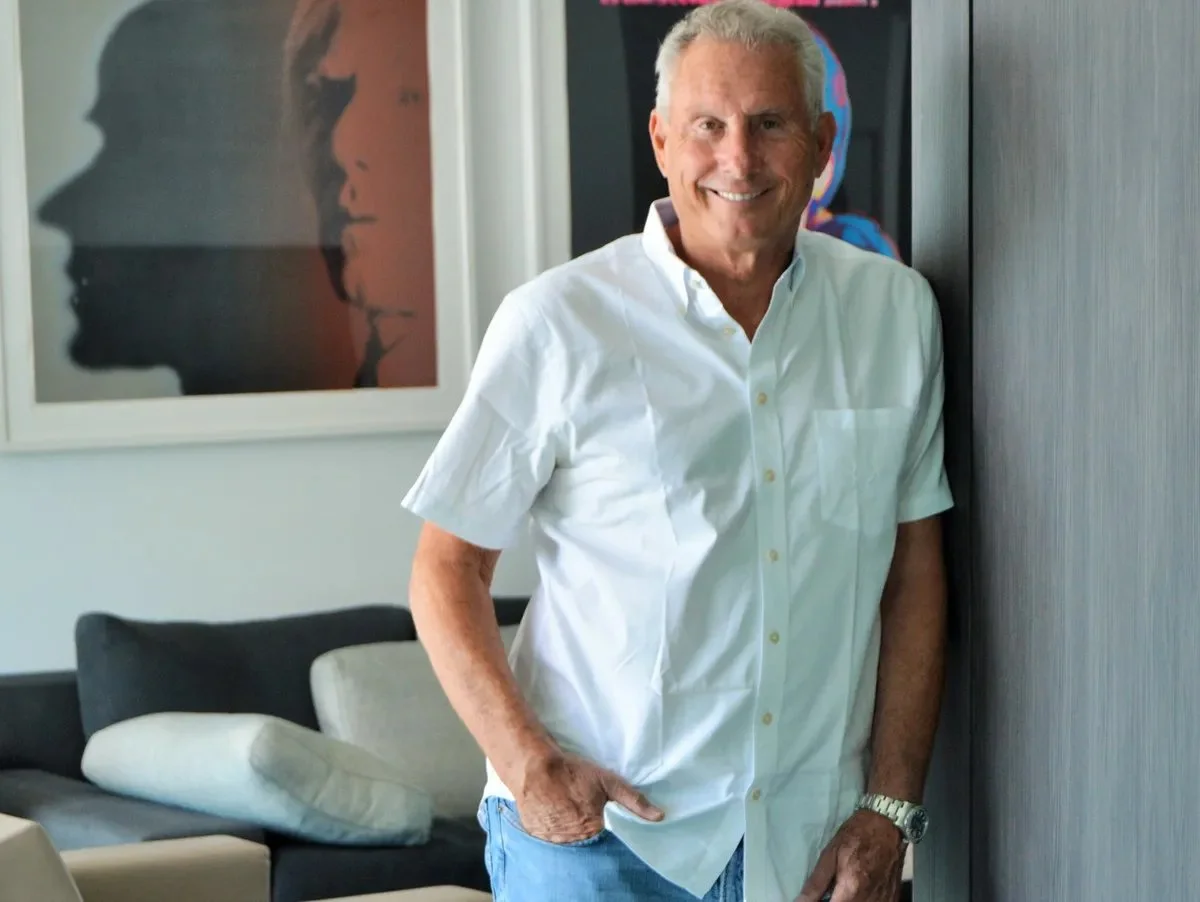 A smiling older man wearing a white short-sleeved shirt and blue jeans stands indoors with one hand in his pocket, next to a textured wall, with a modern sofa with pillows and abstract artwork in the background.