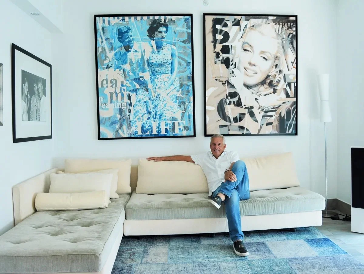 A man sitting on a light-colored modern sofa in a living room with abstract artwork of Marilyn Monroe and another woman hanging behind him.