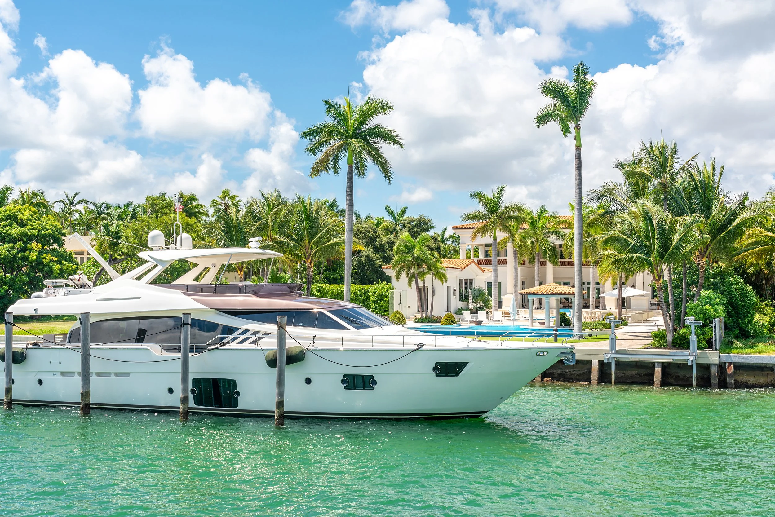 Luxury yacht docked in a tropical marina with palm trees, white buildings, and a bright blue sky with some clouds.
